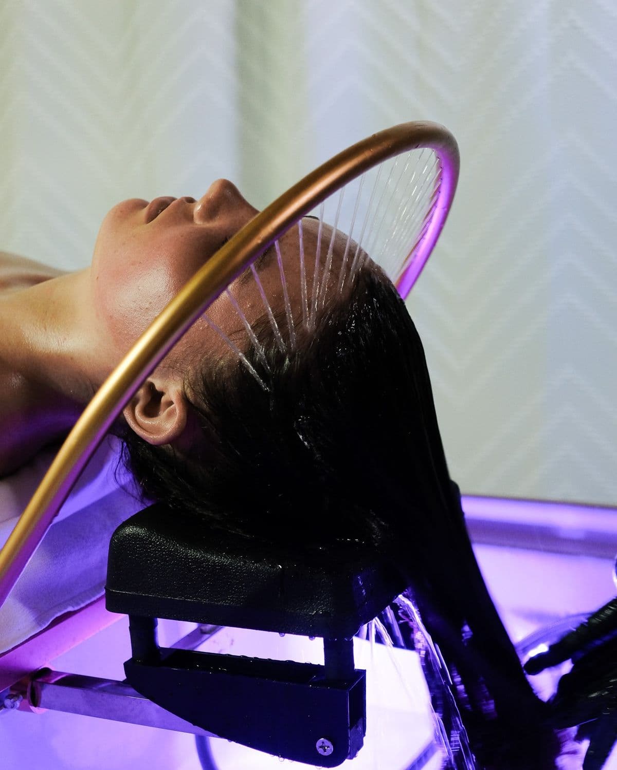 Mujer recibiendo un tratamiento de spa capilar con agua cayendo de un aro dorado.